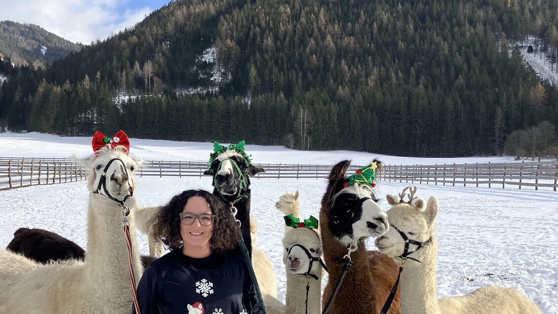 Eine Frau steht im Schnee mit mehreren Alpakas, die festliche Accessoires tragen. Im Hintergrund sind Wälder und Berge zu sehen. | © Alpaka, Lamas und Rentiere zum Grünen See