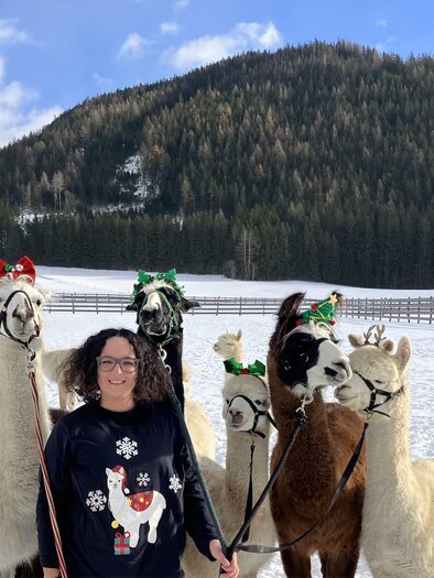 A woman stands in the snow with several alpacas wearing festive accessories. In the background, forests and mountains can be seen. | © Alpaka, Lamas und Rentiere zum Grünen See