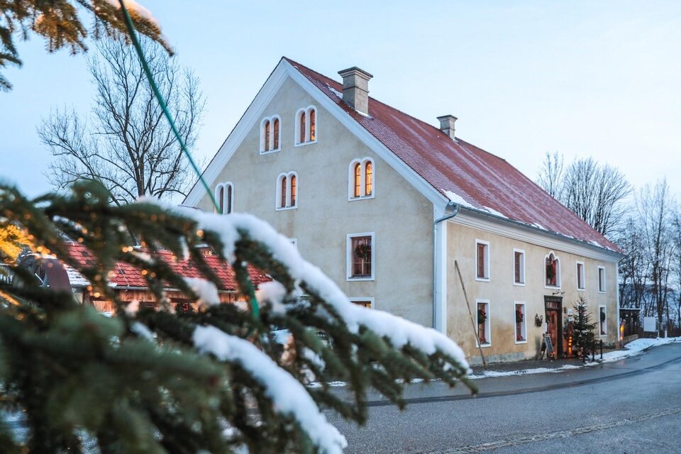 Ein charmantes Haus mit rotem Dach und weißen Wänden, umgeben von schneebedeckten Bäumen. Die Fenster des Hauses strahlen Wärme aus, während die kalte Winterlandschaft im Hintergrund sichtbar ist. | © © Foto De Monte