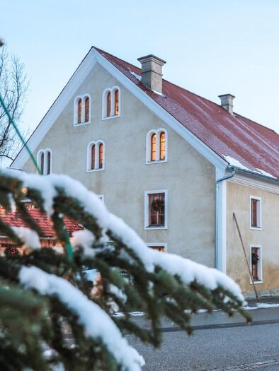 A charming house with a red roof and white walls, surrounded by snow-covered trees. The windows of the house radiate warmth, while the cold winter landscape is visible in the background. | © © Foto De Monte