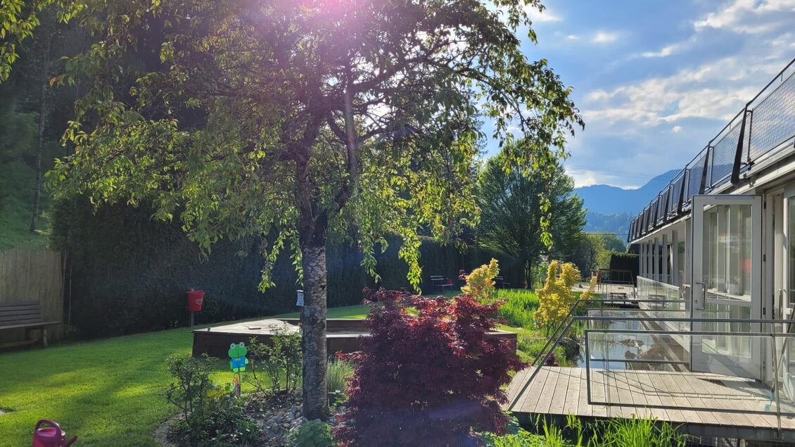 Ein schöner Garten mit üppigem Grün und einem Baum im Vordergrund. Im Hintergrund sind moderne Gebäude und die Berge sichtbar. | © Tim Fuchsberger 