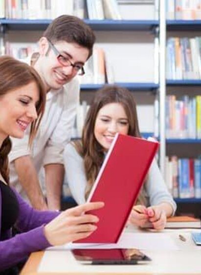 Drei Studierende sitzen an einem Tisch in einer Bibliothek und arbeiten zusammen. Eine der Frauen schaut lächelnd auf ein Buch, während die anderen beiden interessiert zuschauen.