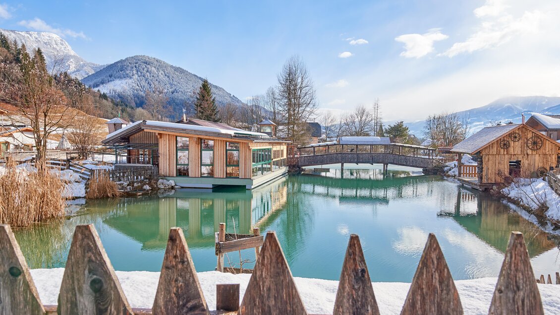 Eine malerische Winterlandschaft mit einem ruhigen See und schneebedeckten Bergen im Hintergrund. Moderne Gebäude und eine Holzbrücke reflektieren sich im klaren Wasser. | © Abenteuerhof Schiefer