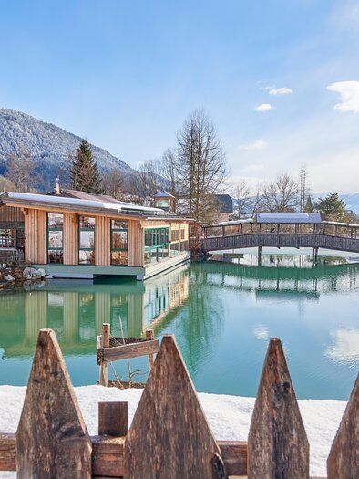 A picturesque winter landscape with a tranquil lake and snow-covered mountains in the background. Modern buildings and a wooden bridge are reflected in the clear water. | © Abenteuerhof Schiefer