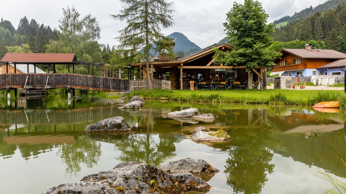 Ein malerischer Teich mit Felsen und Bäumen, umgeben von einer charmanten Holzstruktur. Im Hintergrund sind sanfte Hügel und ein bewölkter Himmel zu sehen. | © Abenteuerhof Schiefer