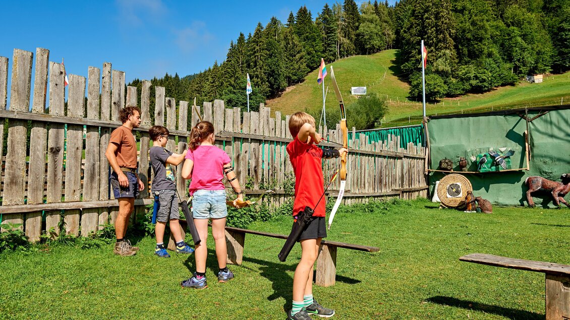 Kinder üben das Bogenschießen auf einer Wiese, umgeben von einem Holzzaun. Im Hintergrund sind Bäume und ein grüner Hügel zu sehen. | © Abenteuerhof Schiefer