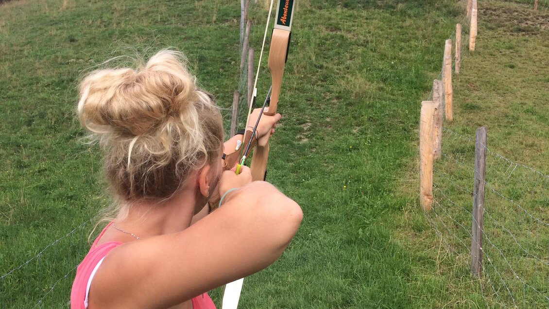 Eine Frau schießt mit einem Bogen auf ein Ziel auf einer Wiese. Im Hintergrund sind Bäume und eine Hügellandschaft zu sehen. | © Familie Reisner