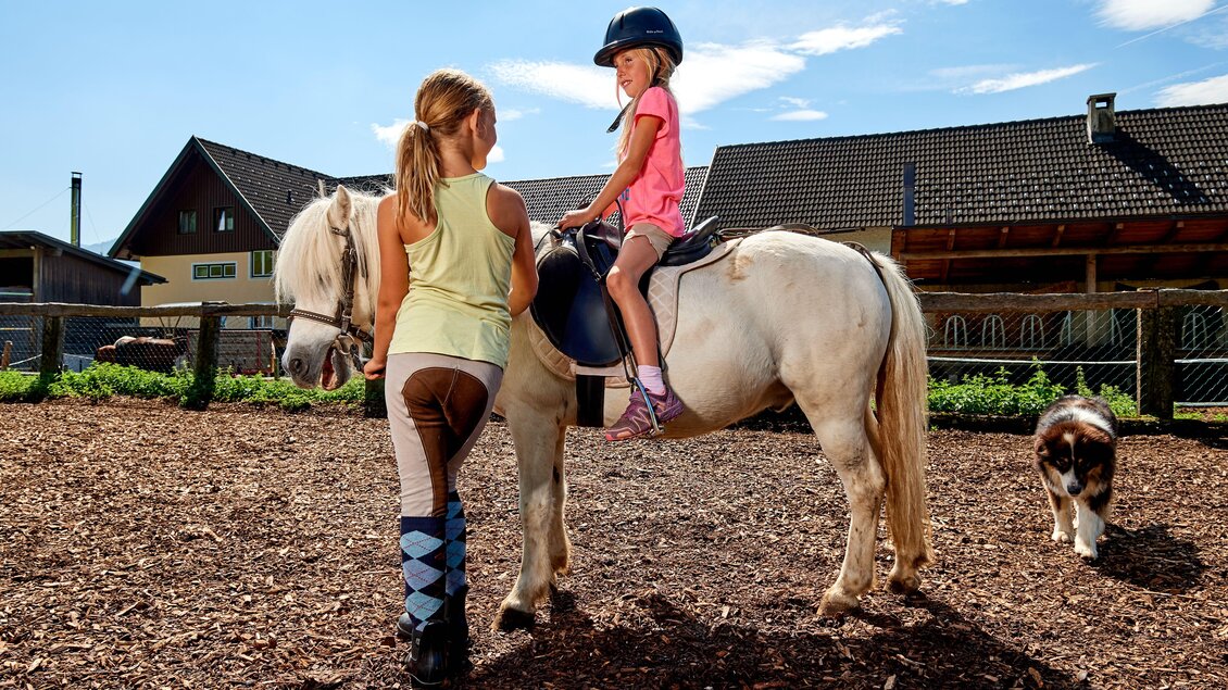Zwei Mädchen sind im Pferdestall. Ein Mädchen sitzt auf einem weißem Pony, während das andere Kind es hält. | © Abenteuerhof Schiefer