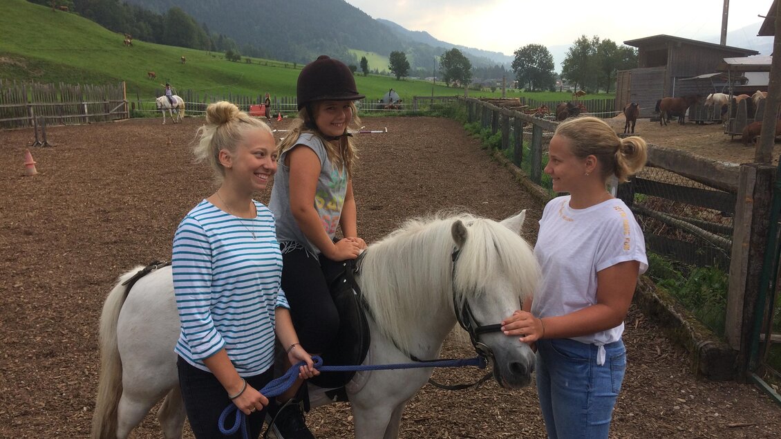 Ein Mädchen sitzt auf einem kleinen Pony, während zwei Frauen daneben stehen. Die Umgebung ist ländlich mit einem Blick auf Berge und eine weite Wiese. | © Familie Reisner