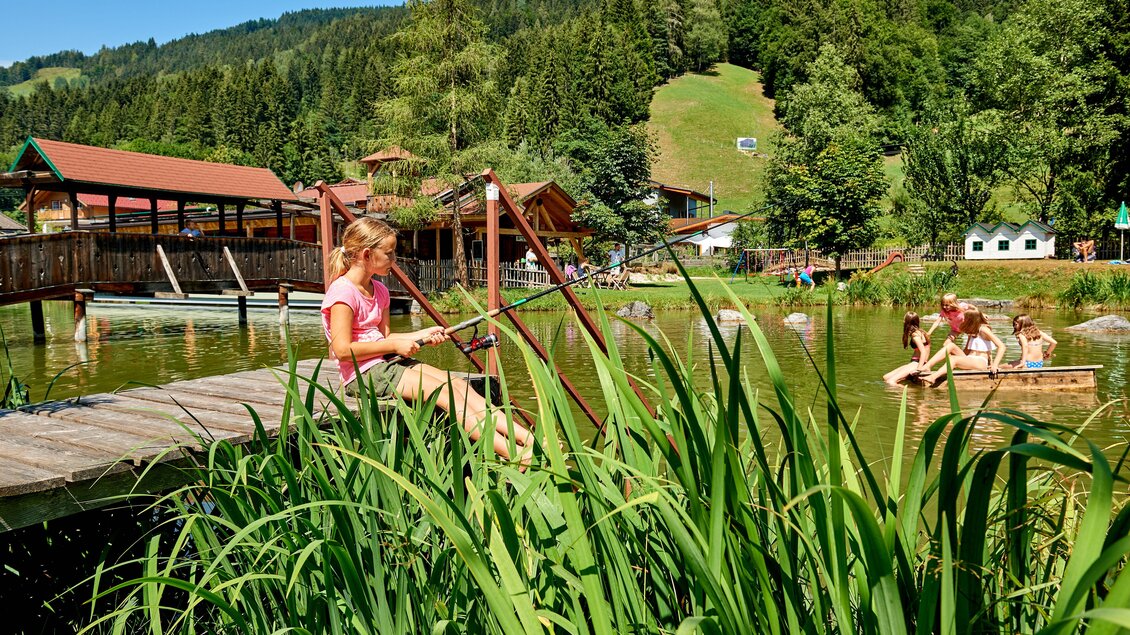 Ein Mädchen sitzt auf einem Steg und angelt an einem ruhigen Teich. Im Hintergrund sind Bäume und spielende Kinder im Wasser sichtbar. | © Abenteuerhof Schiefer