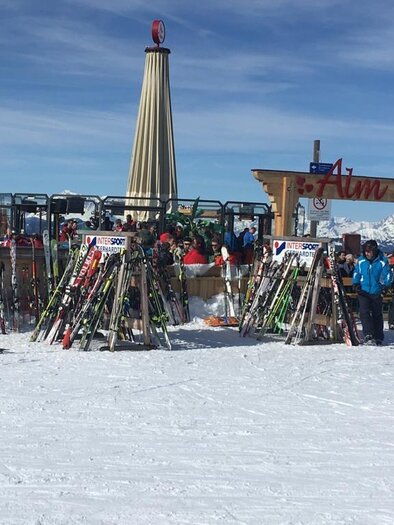 A bustling ski resort with numerous skiers and a mountain hut bar in the background. The surroundings are snow-covered and surrounded by mountains. | © Almbar