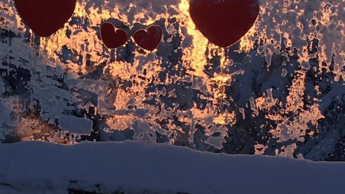 Luftballons in Herzform hängen vor einer verzierten frostigen Fensterscheibe. Im Hintergrund strahlt die Sonne in einem klaren, winterlichen Himmel. | © Almbar