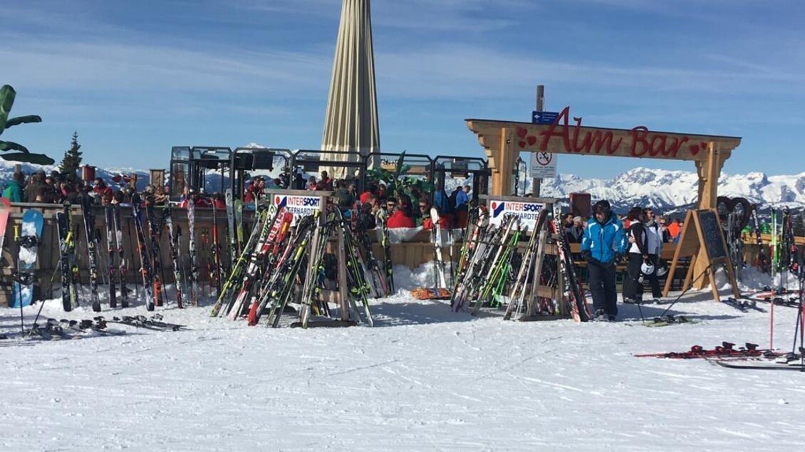 Eine lebhafte Skibar auf einem schneebedeckten Berg mit vielen Skiern und zahlreichen Besuchern. Im Hintergrund sind schneebedeckte Berge und ein blauer Himmel sichtbar. | © Almbar
