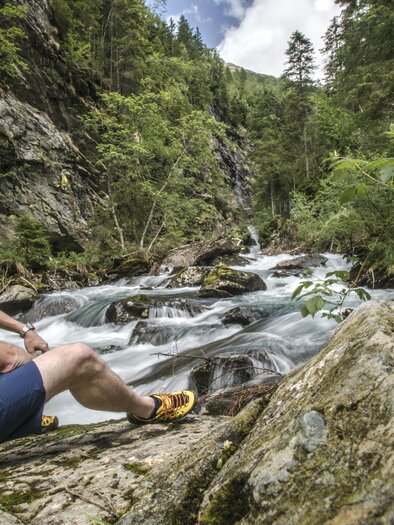 A hiker sits on a rock next to a babbling brook. Surrounded by green trees and boulders, he enjoys the peaceful nature. | © Gerhard Pilz