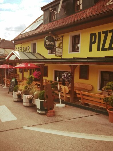 Eine bunte Pizzeria mit sonnigen Terrassen und Pflanzen steht an einer Straße. Rote Sonnenschirme laden zum Verweilen ein. | © Altes Bierhäusl