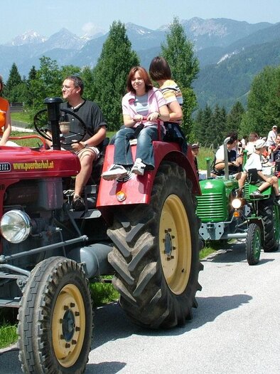 A red tractor with several people is driving on a rural road. In the background, green forests and mountains can be seen. | © Pürcherhof