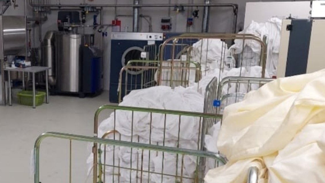 Ein sauberer Waschraum mit mehreren Wäschewagen voller weißer Wäsche. Maschinen und Ausrüstung für die Wäschebearbeitung sind im Hintergrund sichtbar. | © Amadé Textilservice GmbH