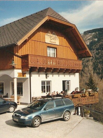 A cozy wooden house in the mountains with a terrace and guests. In front of the house, several cars are parked in a stony area. | © Andreas Höflechner
