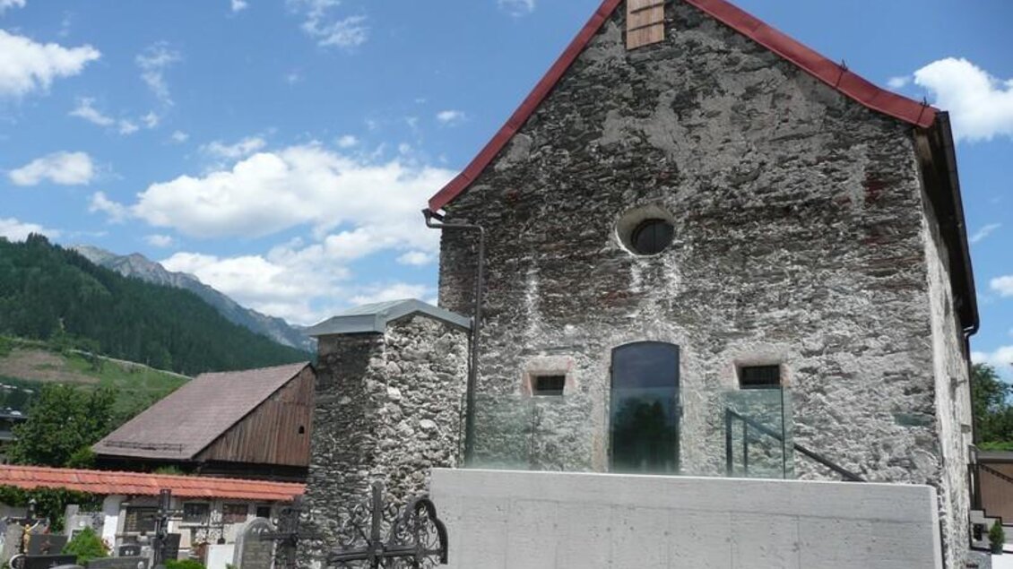 Ein historisches Gebäude mit rotem Dach und Steinmauer steht inmitten eines Friedhofs. Im Hintergrund sind grüne Berge und ein blauer Himmel zu sehen. | © Pfarrverband Schladming