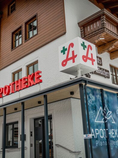 Eine Apotheke mit einem modernen Gebäude im alpinen Stil. Große Schilder deuten auf die Dienstleistungen hin. | © Apotheke Planai West KG