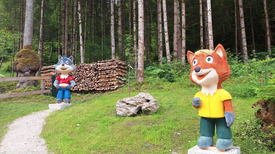 Zwei bunte Figuren eines Wolfes und eines Fuchses stehen in einem Wald. Im Hintergrund sind Holzstapel und Bäume sichtbar. | © Nadine Schrempf