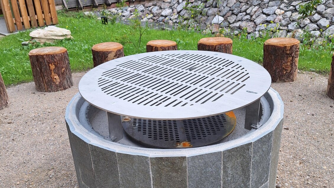 Ein runder Grillplatz aus Stein mit einer Metallabdeckung. Rundherum stehen Holzscheite als Sitzgelegenheiten. | © Nadine Schrempf