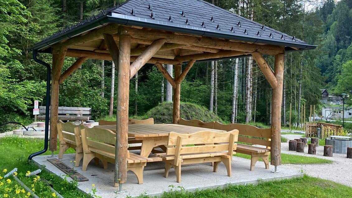 Ein Pavillon mit Holztischen und Bänken steht in einer grünen Umgebung. Im Hintergrund sind hohe Bäume und ein Weg zu sehen. | © Nadine Schrempf