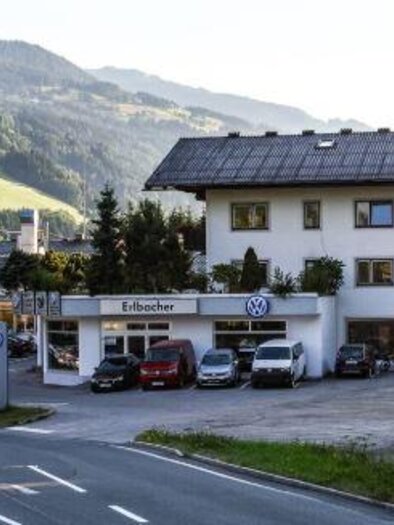 A car dealership with several vehicles in front of a two-story building. In the background, green mountains and a clear sky are visible. | © Autohaus Erlbacher
