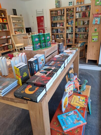 A cozy bookstore with shelves full of books. Various books and children's games are displayed on a wooden table. | © Bücher Geschenke und mehr