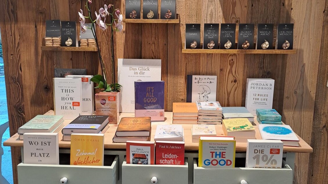 Ein dekorativer Tisch mit verschiedenen Büchern, die in mehreren Farben angeordnet sind. Im Hintergrund sind Regale mit weiteren Produkten und Dekorationen zu sehen. | © Bücher Geschenke und mehr