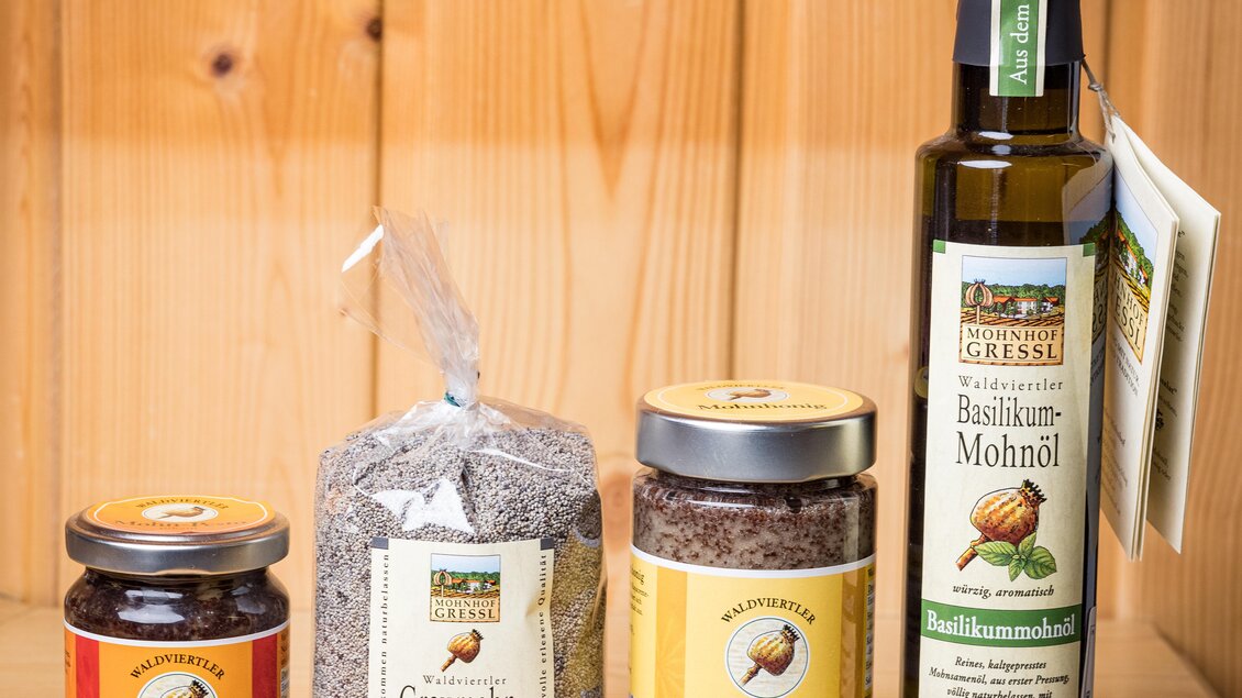 Vier verschiedene Mohnprodukte stehen auf einem Holzregal: Mohn-Pesto, Graumohn, Mohnhonig und Basilikum-Mohnöl. Jedes Produkt hat ein ansprechendes Etikett und ist in Gläsern oder Verpackungen präsentiert. | © Bauernladen Gröbming