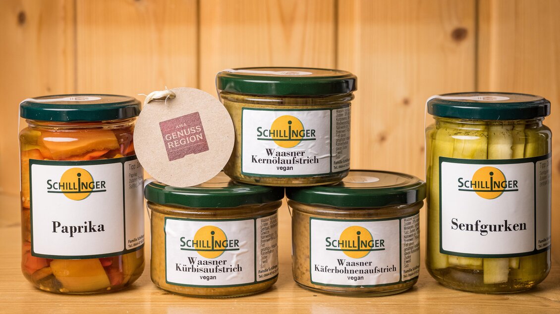 Fünf verschiedene Gläser mit eingelegtem Gemüse auf einem Holzregal. Die Produkte sind von der Marke Schillinger und umfassen Paprika, Kürbis, Keren, und Senfgurken. | © Netzwerk Kulinarik wildbild