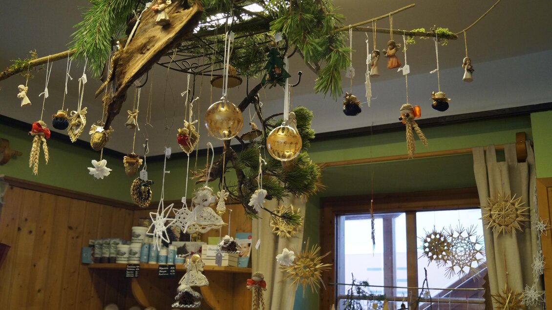 Ein dekorativer Weihnachtsbaum mit verschiedenen Ornamenten und handgemachten Figuren hängt an einem Ast. Im Hintergrund sind weitere Weihnachtsdekorationen und Fenster zu sehen. | © Bauernladen Gröbming