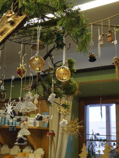 A decorative Christmas tree with various ornaments and handmade figures hangs from a branch. In the background, more Christmas decorations and windows are visible. | © Bauernladen Gröbming