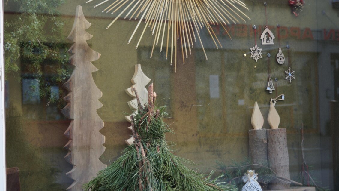 Eine festlich dekorierte Schaufensterfront mit einer großen goldenen Sonne aus Stroh und handgemachten Holzfiguren. Im Vordergrund steht ein schöner Tannenbaum, der fröhliche Weihnachtsstimmung vermittelt. | © Bauernladen Gröbming