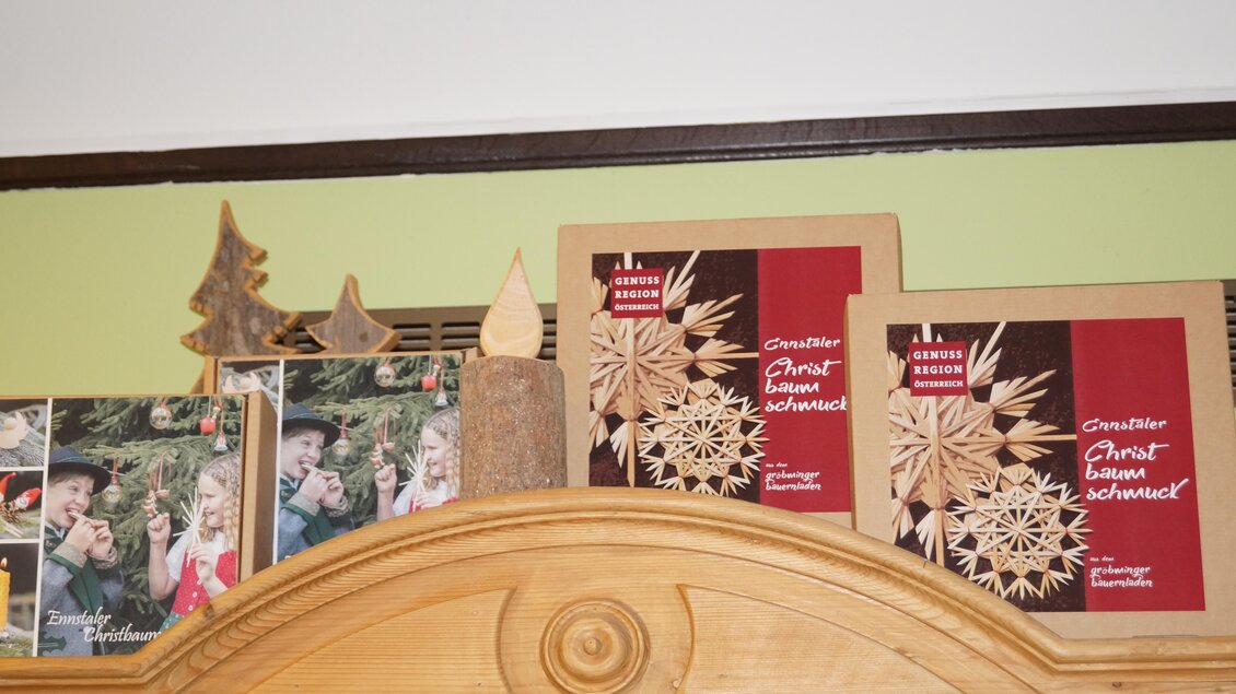 Eine dekorative Holzablage mit Weihnachtsornamenten. Darunter befinden sich zwei Bilder von Personen und zwei Verpackungen mit Weihnachtsdekoration. | © Bauernladen Gröbming