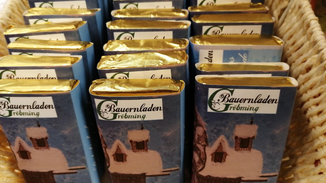 Eine Korb mit mehreren Schokoladenriegeln in blauer Verpackung. Die Riegel sind mit einer winterlichen Hütte abgebildet und haben goldene Folie oben. | © Marianne Ritzinger 