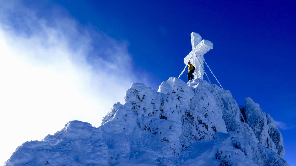 Ein Berggipfel mit einer großen, schneebedeckten Kreuz. Eine Person steht oben und genießt die Aussicht unter blauem Himmel. | © Heli Rettensteiner
