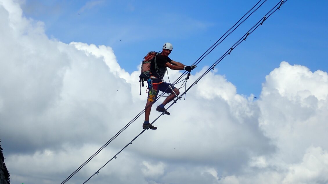 Ein Kletterer balanciert auf Drahtseilen inmitten einer beeindruckenden Wolkenlandschaft. Im Hintergrund sind Berge und ein klarer Himmel sichtbar. | © Monte-Krah