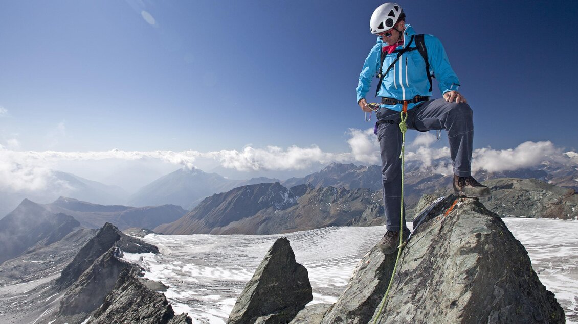 Ein Bergsteiger steht auf einem Felsen und schaut auf die umliegende Berglandschaft. Der Himmel ist klar und es sind einige Wolken zu sehen. | © Monte-Krah