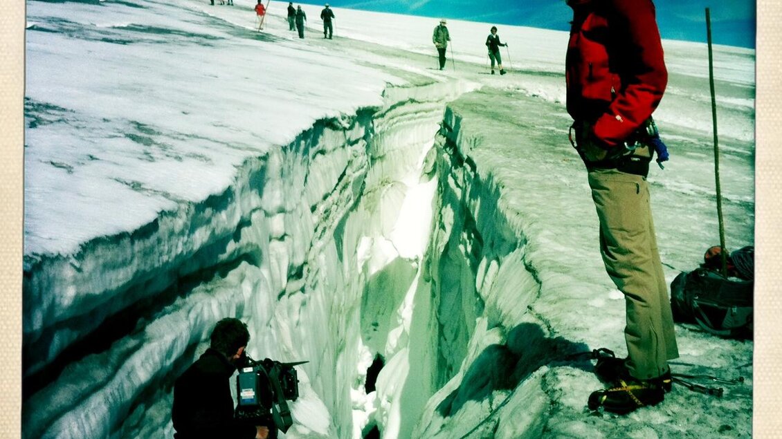 Eine Gruppe von Menschen steht auf einem schneebedeckten Berg. Im Vordergrund filmt jemand mit einer Kamera eine tiefe Spalte im Eis. | © Monte-Krah