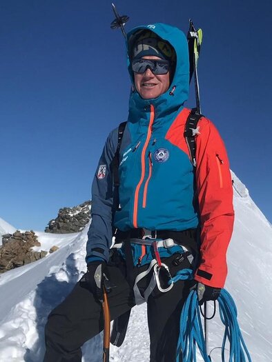 A mountaineer stands on a snow-covered peak. The sky is clear and blue, and he is wearing a colorful winter jacket.