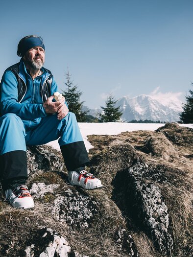 A man is sitting on a rock in the mountains and enjoying the view. In the background, there are snow-covered peaks and green trees. | © Anton Kerschbaumer