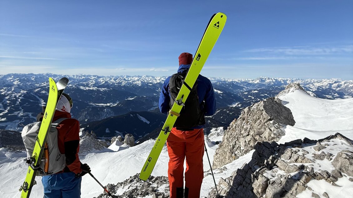 Zwei Skifahrer stehen auf einem schneebedeckten Gipfel und blicken auf die Berge. Der Himmel ist klar und blau. | © Michael Perhab