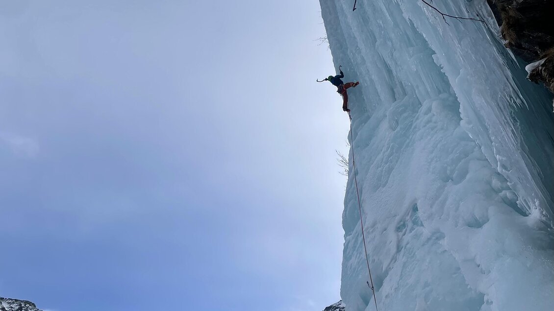 Ein Eiskletterer erklimmt einen gefrorenen Wasserfall in einer bergigen Landschaft. Der Himmel ist bewölkt und die Umgebung ist von Schnee bedeckt. | © Michael Perhab