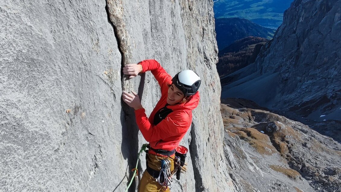 Ein Kletterer in leuchtend roter Kleidung steigt eine steile Felswand hinauf. Im Hintergrund sind Berge und ein klarer Himmel zu sehen. | © Michael Perhab