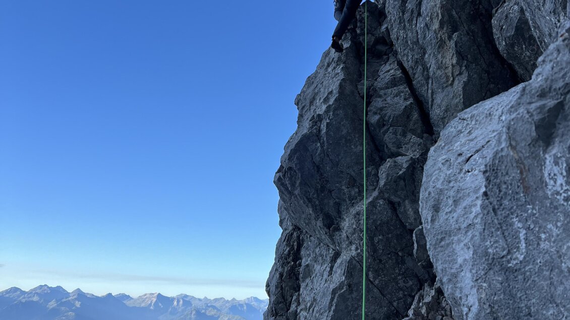 Ein Bergsteiger klettert an einer steilen Felswand. Im Hintergrund sind schneebedeckte Berge und ein klarer blauer Himmel zu sehen. | © Michael Perhab