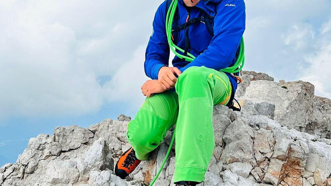 Ein Kletterer sitzt auf einem Felsen und lächelt. Er trägt eine Schutzbrille und eine leuchtend grüne Kletterausrüstung. | © Michael Perhab