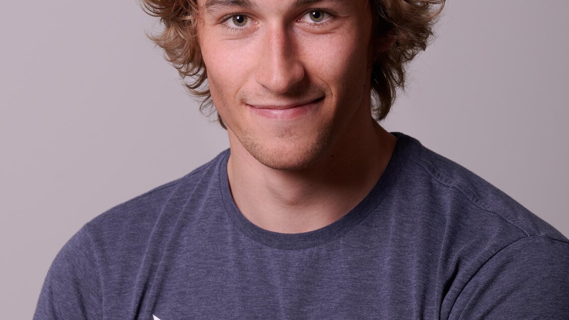 Ein junger Mann mit lockigem Haar lächelt in die Kamera. Er trägt ein blaues T-Shirt mit einem Logo. | © Michael Perhab