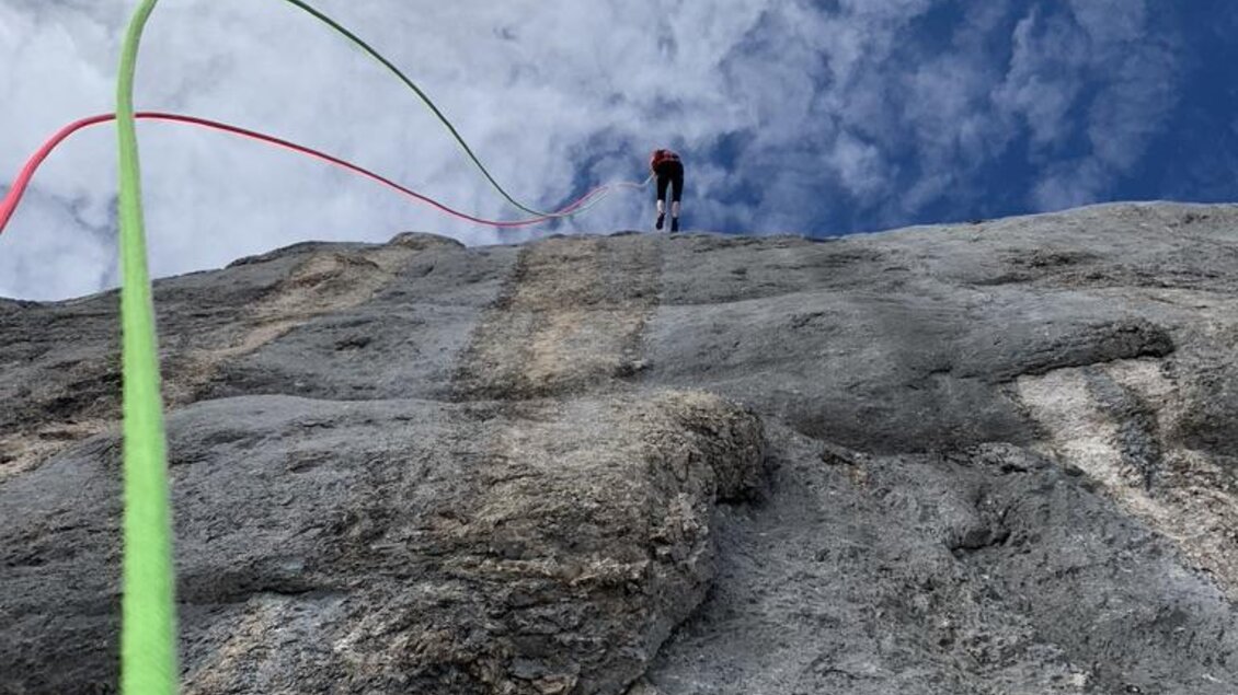 Ein Kletterer steht auf einer Felswand unter einem blauen Himmel mit Wolken. Bunte Kletterseile verlaufen von oben nach unten. | © Peter Perhab